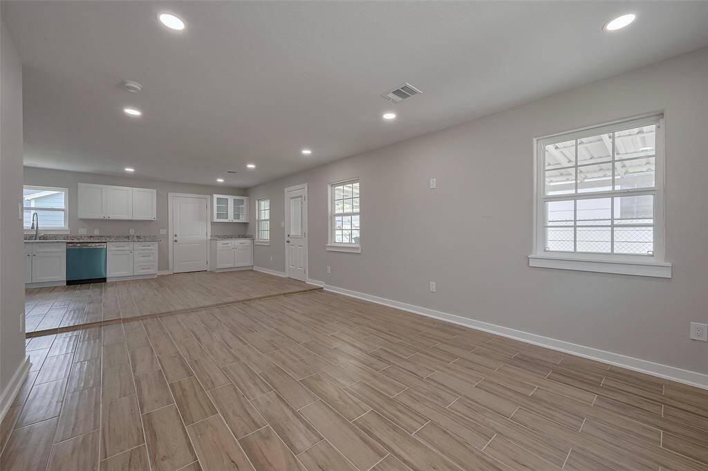 Home shows an open-concept living area with light wood-look tile flooring and neutral walls. The space leads into a kitchen with white cabinetry, granite countertops, and stainless steel appliances. Recessed lighting and windows brighten the room, creating a modern, airy atmosphere.