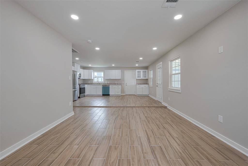 This photo showcases a bright, open-concept living area with wood-look tile flooring and recessed lighting, leading to a modern kitchen equipped with stainless steel appliances and white cabinetry.