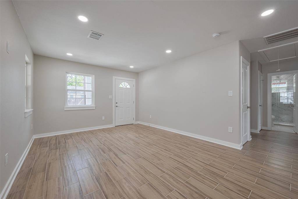 This photo shows a bright, spacious room with light-colored walls and wood-look tile flooring. It features recessed lighting, a window, a white door, and an open doorway leading to another area of the house.