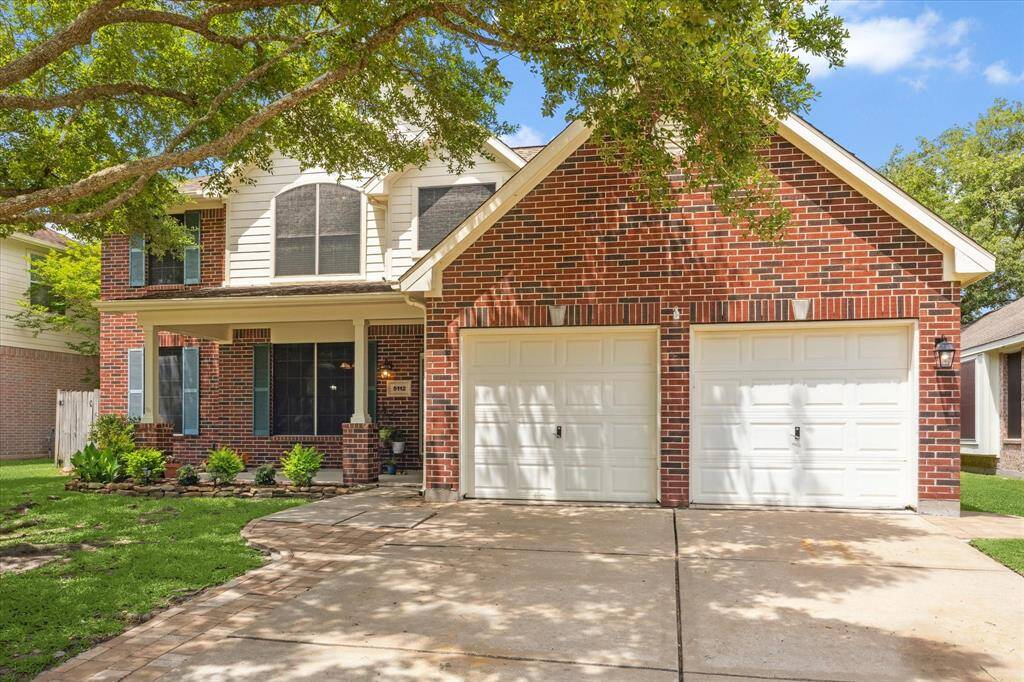 This charming two-story home features a classic red brick exterior, a double garage, and a welcoming front porch. It is surrounded by a well-maintained lawn with mature trees providing shade. Perfect for a family seeking a comfortable and inviting residence.