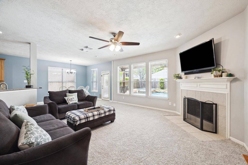 Bright and spacious living room with a cozy fireplace, ceiling fan, and large windows offering plenty of natural light. The open layout includes a view to the backyard and pool creating an inviting atmosphere for family gatherings.