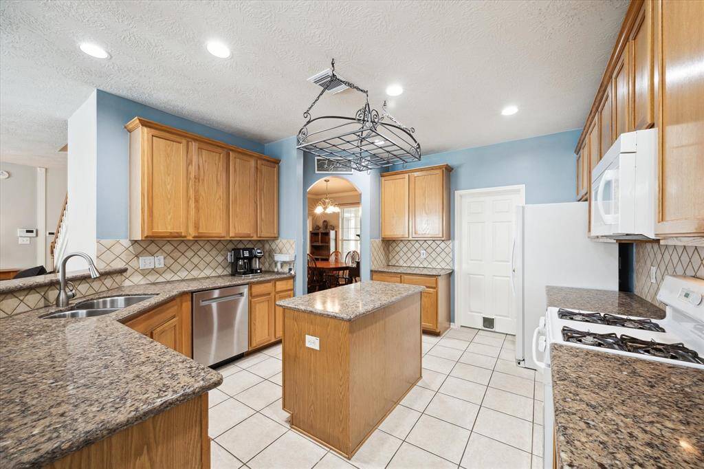 This kitchen features granite countertops, ample wooden cabinetry. It includes a central island, a gas stove, a double sink, and a decorative ceiling rack. The space is well-lit with recessed lighting and has a tiled backsplash and floor.