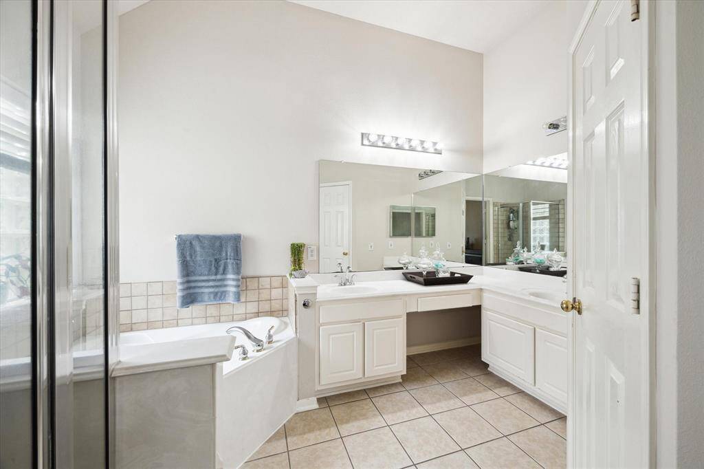 This bathroom features a spacious layout with dual sinks, a large mirror, and ample lighting. It includes a bathtub with tiled surround and a separate shower. The neutral tile flooring and white cabinetry offer a clean, modern look.