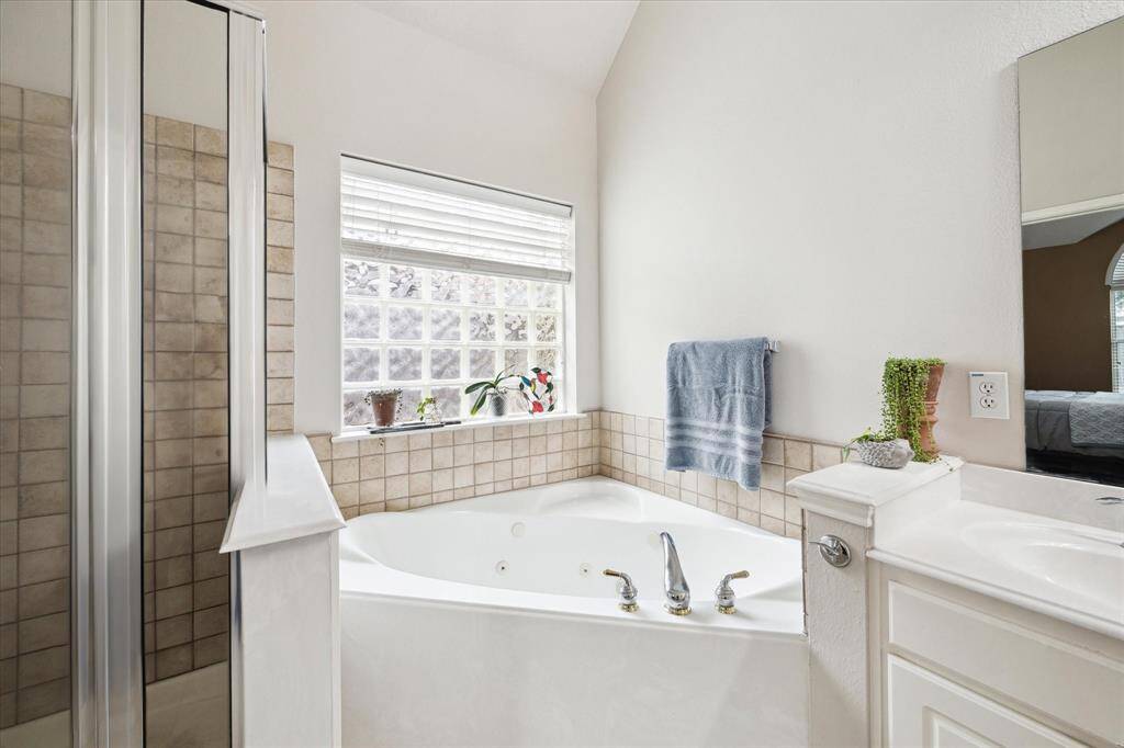 This bathroom features a cozy corner soaking tub surrounded by light-colored tiles. A large window provides natural light, and there’s a separate shower with a glass door. The space is bright and inviting,