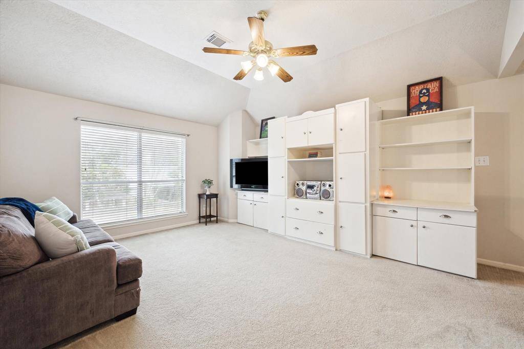 This spacious, carpeted living area features  large window for natural light, ceiling fan, and built-in white shelving with ample storage, perfect for entertainment and relaxation.