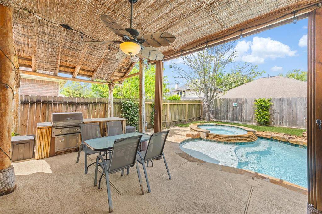 This backyard oasis features a cozy covered patio with a ceiling fan, Free standing grill for outdoor cooking, a dining area, and a beautiful pool with a spa, all surrounded by a privacy fence and lush greenery. Perfect for relaxation and entertaining.
