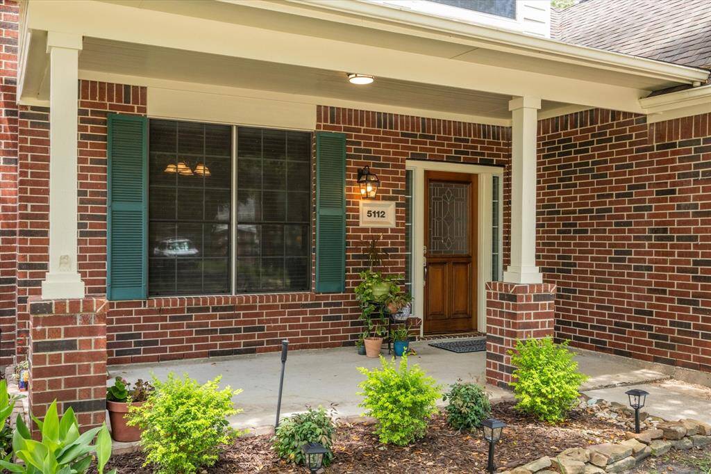 This photo showcases a welcoming front porch with a brick exterior, featuring a wooden door with glass panels, green shutters, The porch is accented by columns and outdoor lighting, creating a charming entrance.