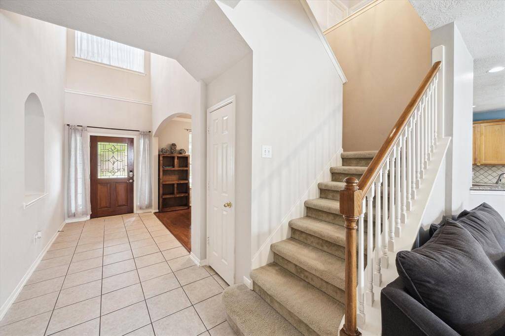 This home features a bright entryway with high ceilings, tiled flooring, and a wooden front door with glass detailing. A carpeted staircase with wooden railing leads to the upper floor, while an archway opens into the living area.