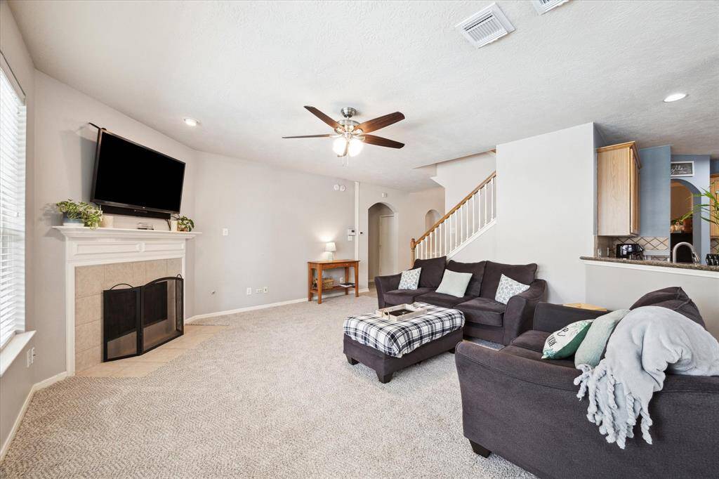 This inviting living room features plush carpet, a cozy fireplace, and a ceiling fan. The adjacent kitchen and staircase add functionality and charm to the space.