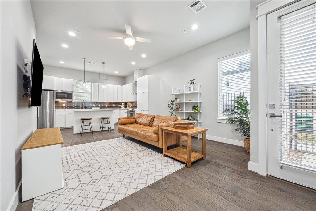 This modern living area features an open-concept layout with a sleek kitchen, complete with stainless steel appliances and a breakfast bar. The space is bright and airy, with large windows, a cozy leather sofa, and stylish wood flooring. Perfect for entertaining or relaxing.