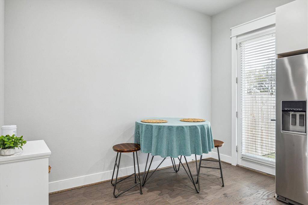 This cozy breakfast nook features a small round table with two stools, set against a bright, minimalist backdrop. Natural light floods in through a door with blinds, adjacent to a sleek stainless steel refrigerator.