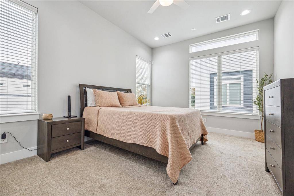 This bright and inviting bedroom features large windows for ample natural light, neutral walls, and a cozy carpet. It includes a modern bed with matching nightstands and dresser, creating a comfortable and stylish space.