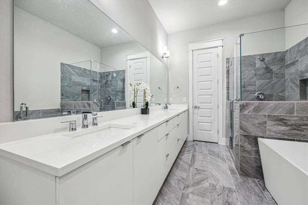 This modern bathroom features a spacious double vanity with sleek white countertops, a large mirror, and elegant fixtures. The walk-in shower has glass doors and stylish gray tile work, complemented by a separate bathtub. The neutral color palette enhances the room's contemporary feel.