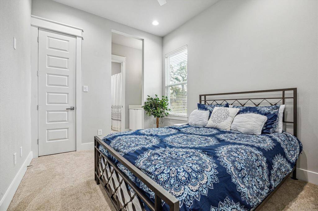 This cozy bedroom features neutral walls, carpet flooring, and ample natural light from a large window. It includes a stylish bed with a decorative blue and white comforter, and a small plant adds a touch of greenery.