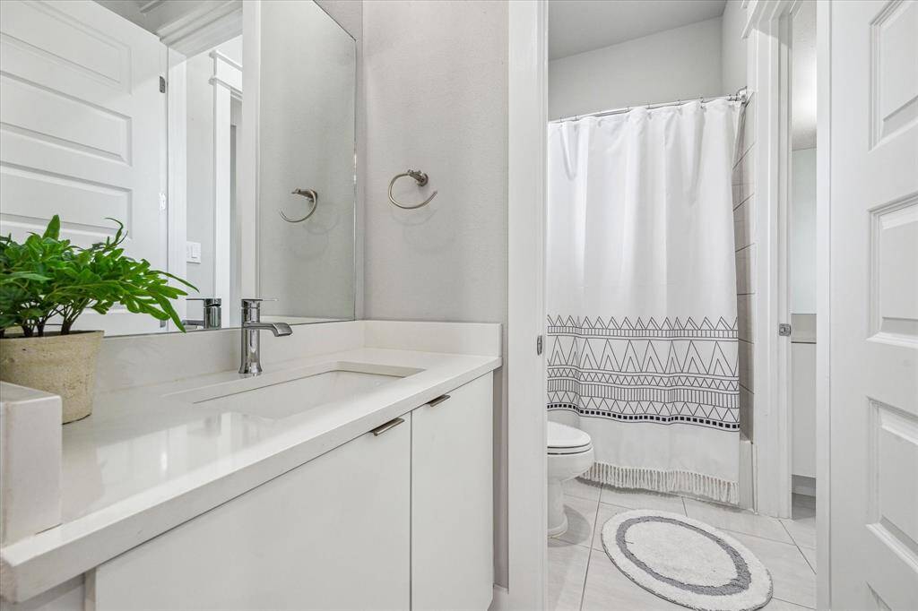 This is a modern, bright bathroom featuring a sleek white vanity, a large mirror, and a shower with a stylish patterned curtain. It has a clean, minimalist design, enhanced by a potted plant for a touch of greenery.