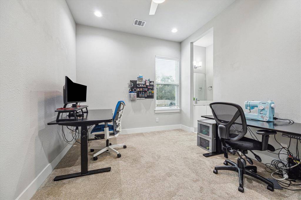 This room is a versatile workspace featuring two desks, a sewing machine, and a computer setup. It has carpeted flooring, soft gray walls, recessed lighting, and a window providing natural light. There's an adjacent bathroom with modern fixtures.