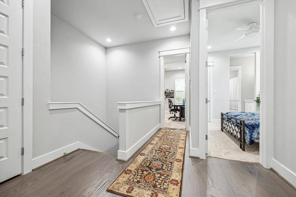 This photo showcases a bright, modern hallway with hardwood floors and recessed lighting. It features a stairway, a patterned runner, and leads to a cozy bedroom and a home office space.