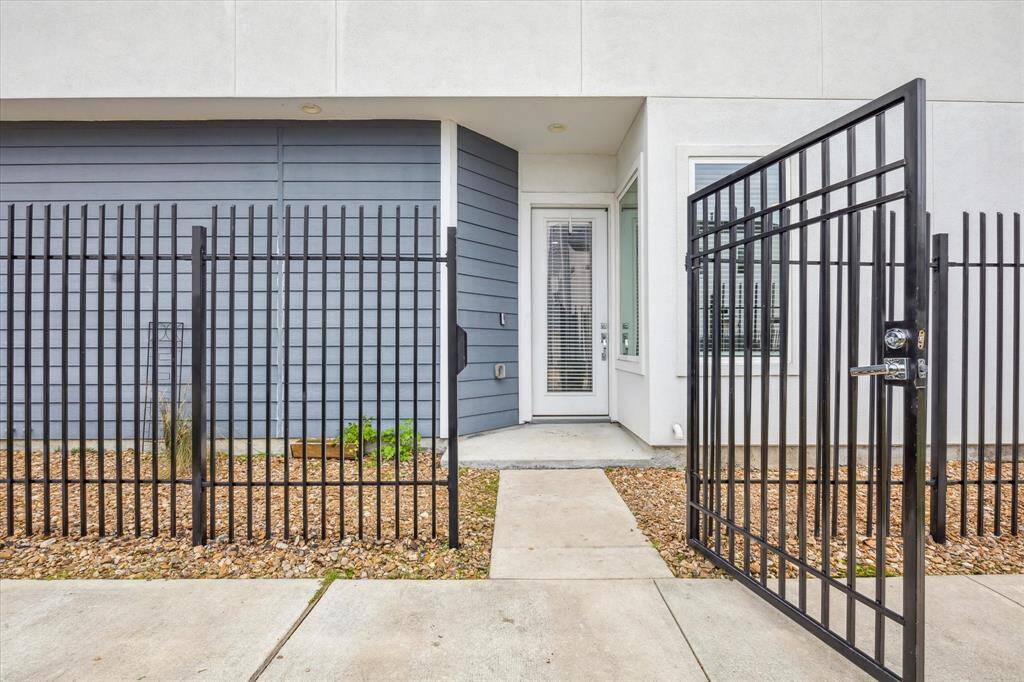 This photo shows a modern entrance with a black metal gate leading to a front door. The exterior features a combination of blue siding and white walls, with a small landscaped area of rocks and plants.