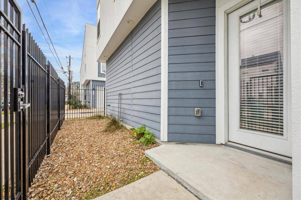 This photo shows a side entrance of a modern home with a glass door and blinds, leading to a gravel pathway. The exterior features blue-gray siding and is enclosed by a black metal fence, offering a neat and secure entryway.