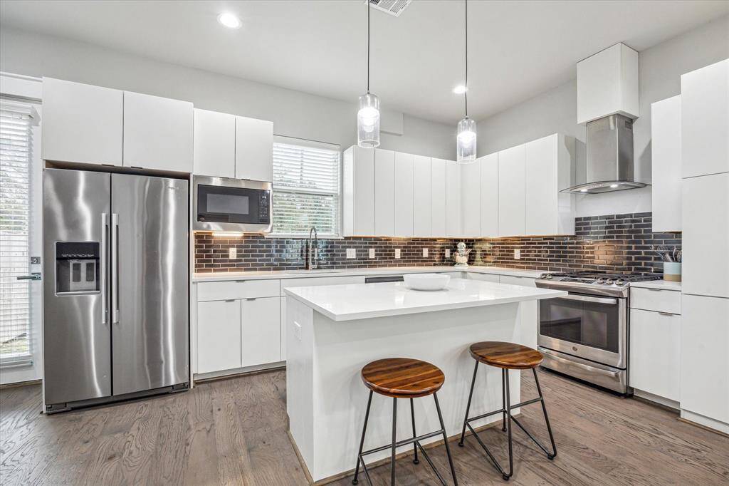 This modern kitchen features sleek white cabinetry, stainless steel appliances, and a stylish dark tile backsplash. It includes a central island with seating and pendant lighting, offering a bright and functional space for cooking and entertaining.