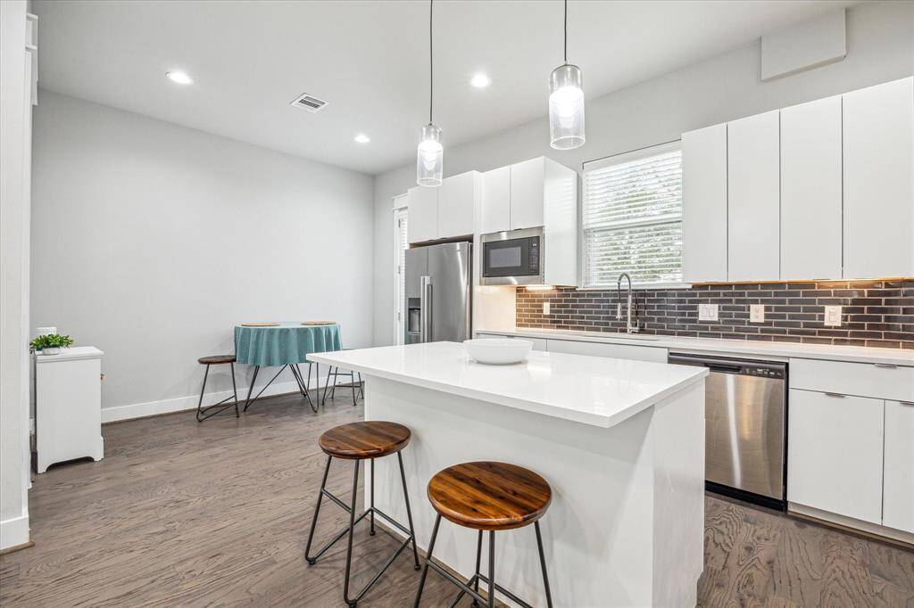 This modern kitchen features sleek white cabinets, a stylish black tile backsplash, and stainless steel appliances. It includes a central island with wooden stools and pendant lighting, complemented by hardwood floors and a cozy breakfast nook.