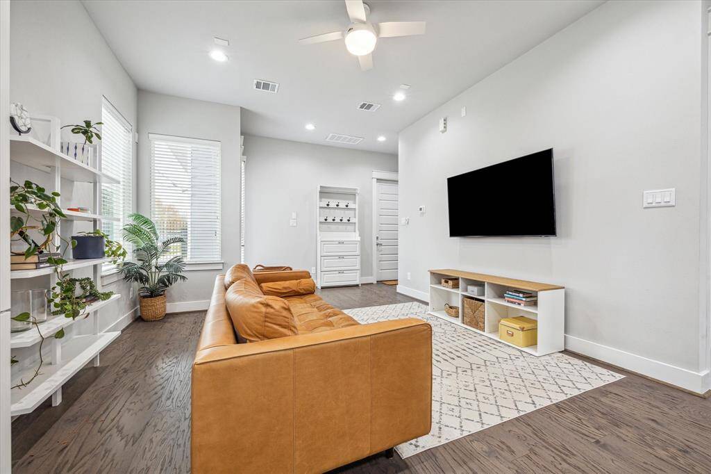 This modern living room features a cozy tan sofa, sleek wall-mounted TV, and stylish shelving with plants. It has wood flooring, a ceiling fan, and ample natural light from the large windows, creating a bright and inviting space.