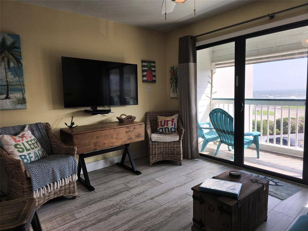 Lovely living area with beachy cool decor, featuring a mounted TV, wicker chairs, and a rustic wooden chest. Large sliding glass doors open to a balcony with Gulf views, creating a bright and inviting space.