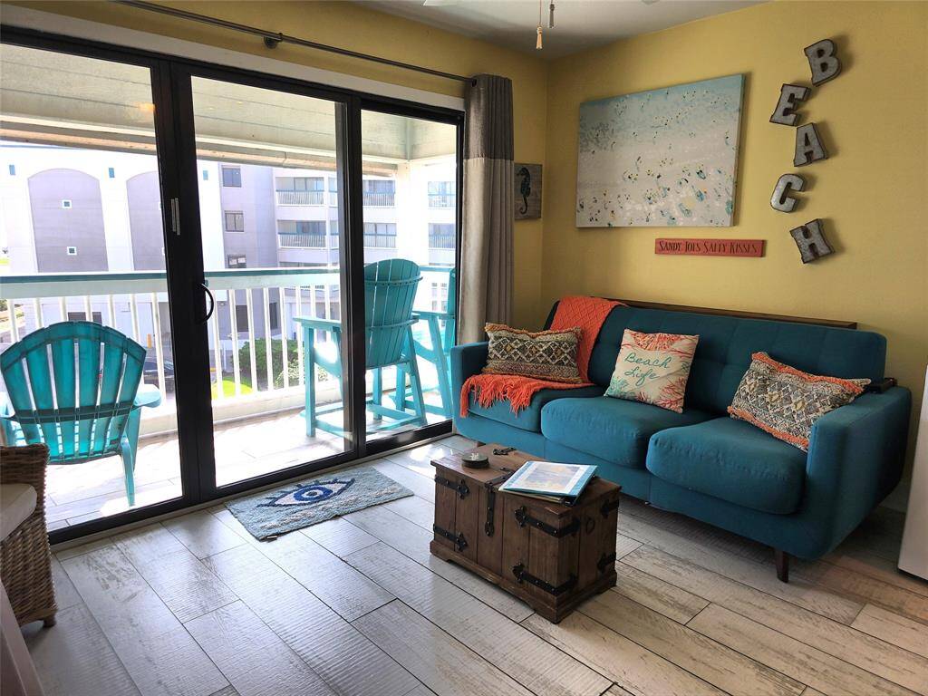 The living room features beach cool decor with a vibrant blue couch and colorful cushions. Sliding glass doors open to a balcony with seating, offering a bright and airy atmosphere. Perfect for a relaxed coastal lifestyle.
