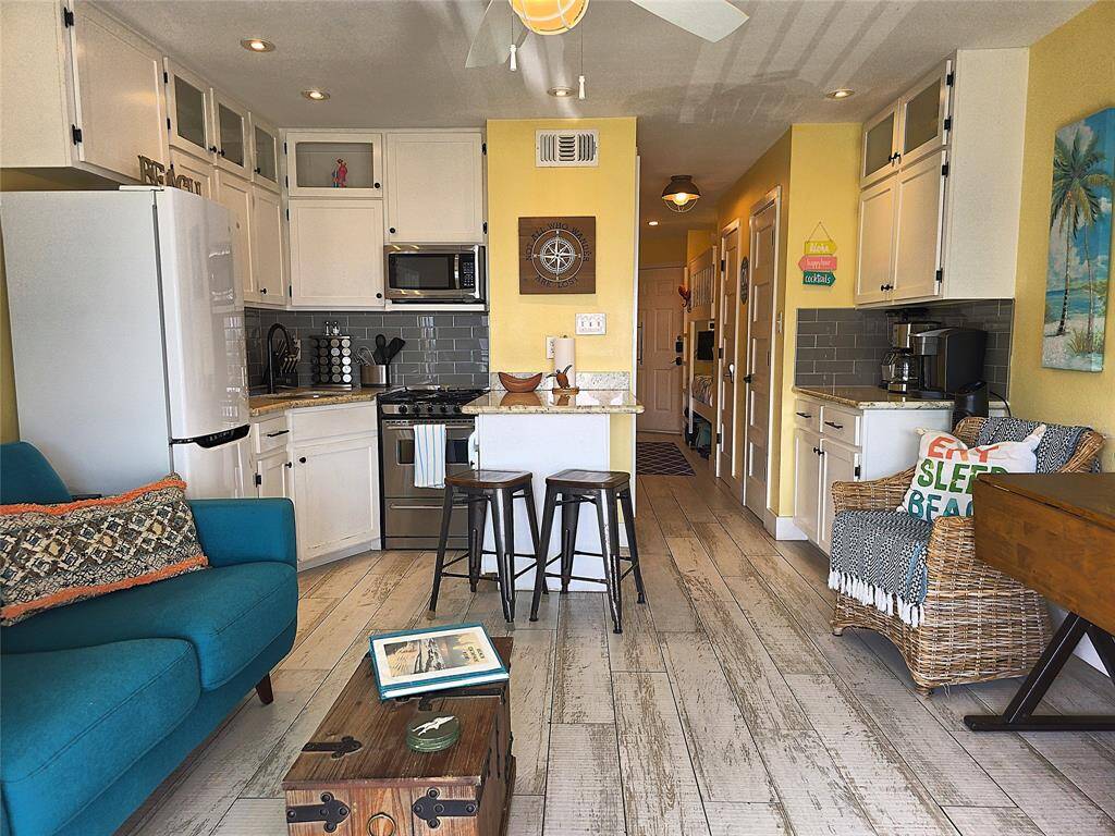 This warm and inviting living space features an open kitchen with white cabinets, modern appliances, and a small breakfast bar. The room is bright with yellow accents, rustic wood flooring, and comfortable seating, creating a charming, beachy atmosphere.