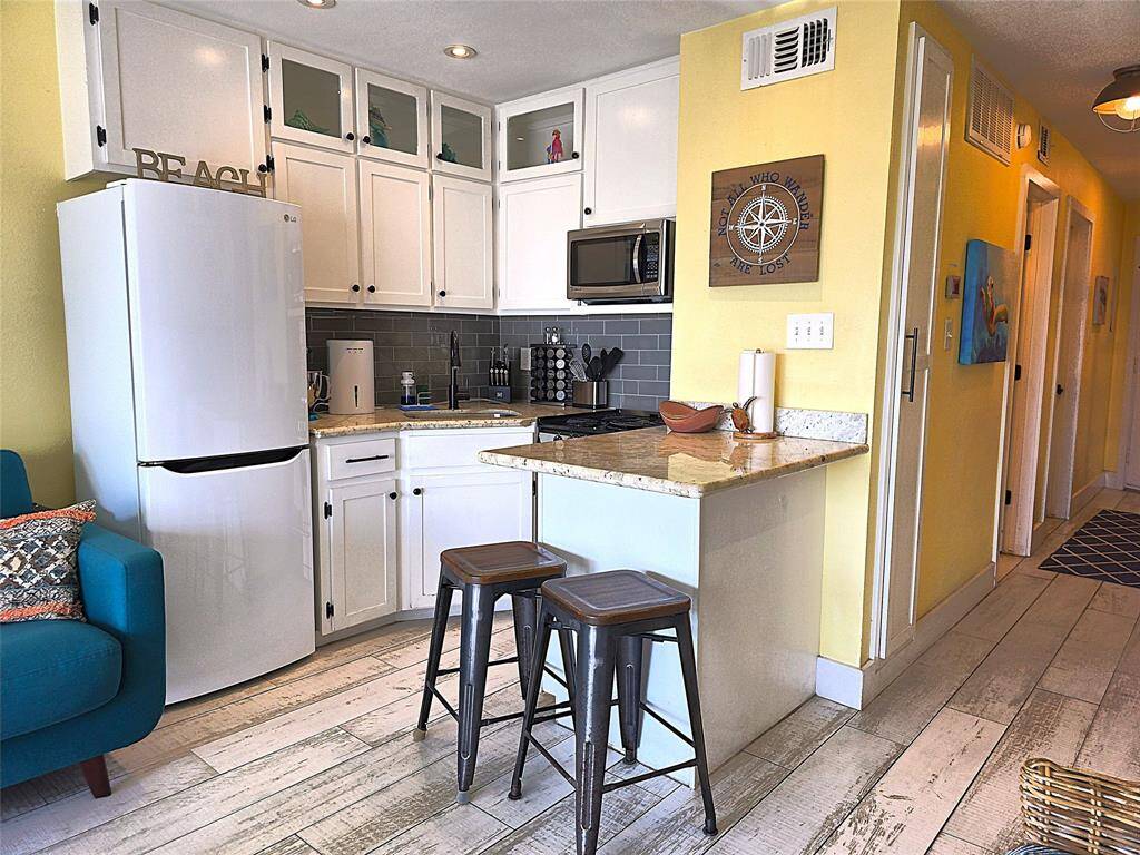 The kitchen has white cabinets, granite countertops, and a breakfast bar with two stools. The kitchen features a modern refrigerator and microwave, with a light grey glass tile backsplash adding sparkle and bling. The open layout leads to a hallway, and the space is well-lit with recessed lighting, creating a warm and inviting atmosphere.