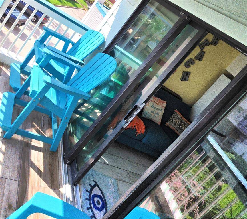Balcony with bright turquoise Adirondack chairs, perfect for relaxing. Through the sliding glass doors, a comfortable living area is visible, decorated with beach-themed accents, creating a vibrant and inviting space.