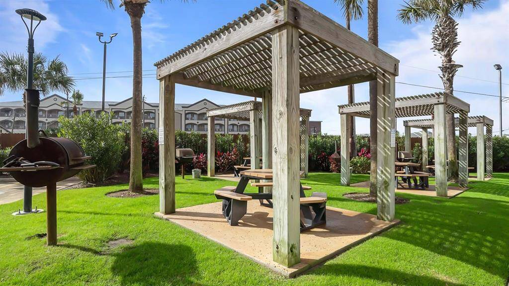 Outdoor BBQ and eating area with wooden pergolas and picnic tables, ideal for gatherings. It features lush green grass, palm trees, and built-in grills, offering a perfect setting for relaxation and entertainment.