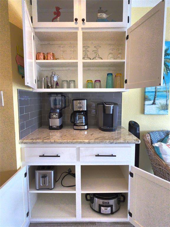The Coffee to Cocktails counter featuring white cabinetry with open shelves displaying glassware. The countertop holds a coffee maker, blender, and a Keurig machine. Below, there's storage for a toaster and a slow cooker. The backsplash is a sleek gray tile.