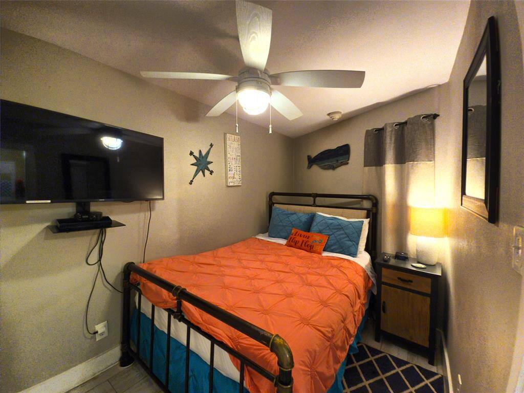 Cozy bedroom with a modern ceiling fan, mounted TV, and nautical decor. Features a comfortable bed with vibrant bedding, a nightstand with a lamp, and a stylish rug. Perfect for a relaxing retreat.