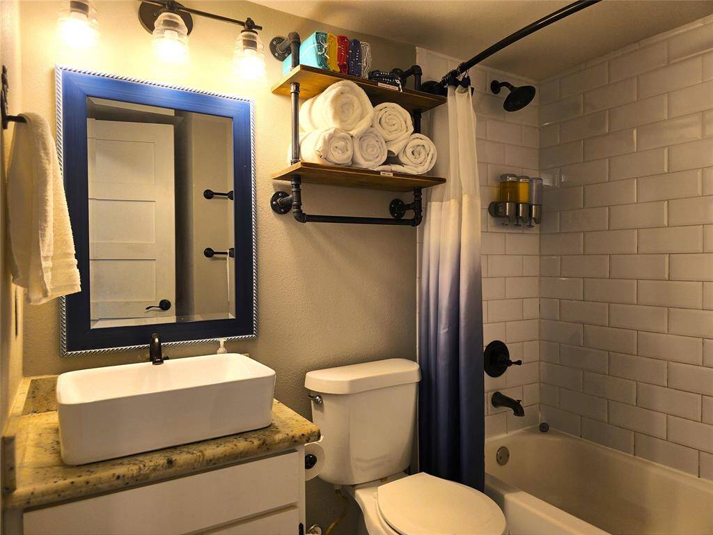 This bathroom features a modern, cozy design with a vessel sink on a granite countertop, a large mirror with a blue frame, and warm lighting. It includes a bathtub with white subway tile, a shower curtain, and black fixtures. Open shelving provides storage with neatly rolled towels.