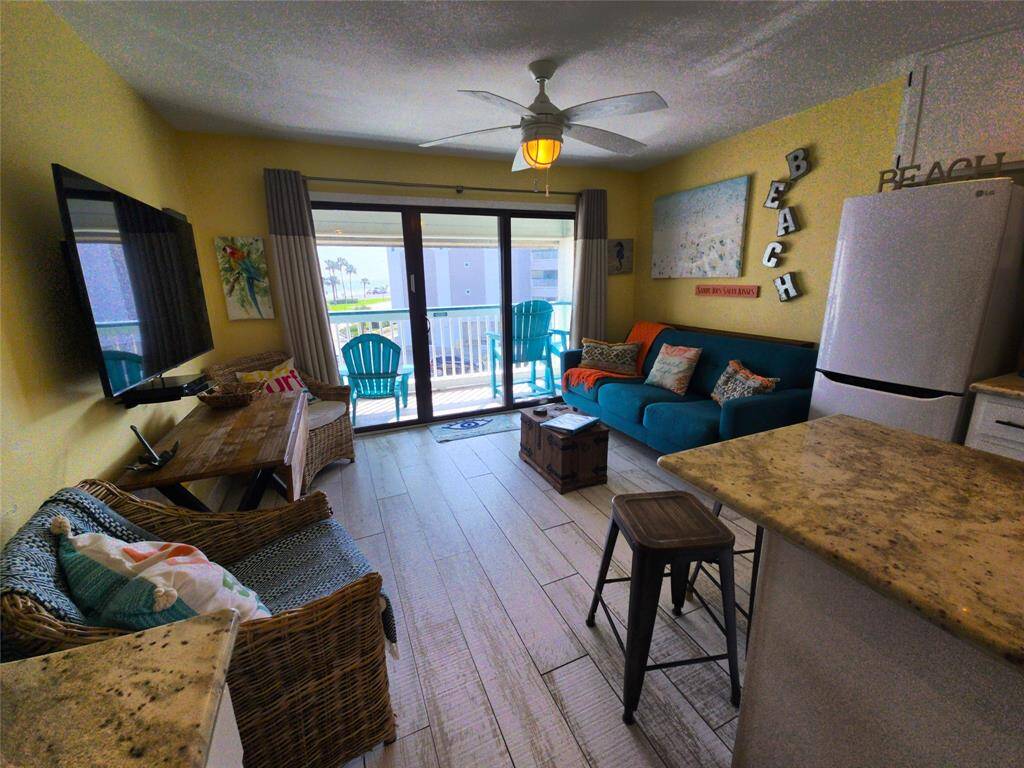 This multipurpose living space features bright and beachy decor with a comfortable sofa, wicker chairs, and a wall-mounted TV. There's a dining area, a kitchen with a breakfast bar, and large glass slider leading to a balcony with Gulf views, perfect for relaxation.