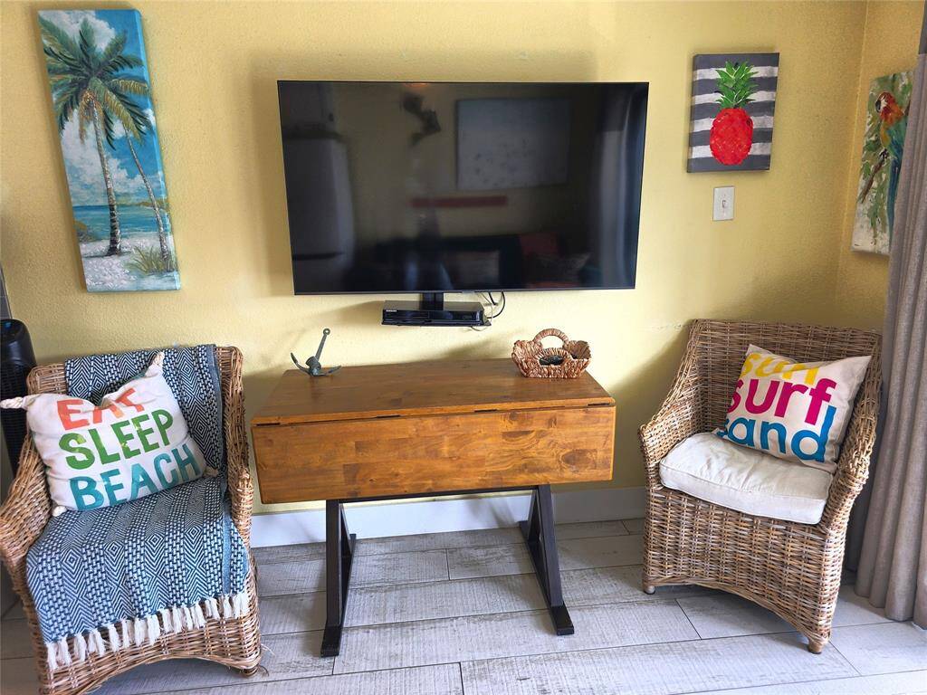 This stylish living area features a mounted TV, wicker chairs with colorful beach-themed pillows, and a wooden drop-leaf table. Vibrant artwork adds a tropical touch, creating a relaxed, inviting atmosphere.