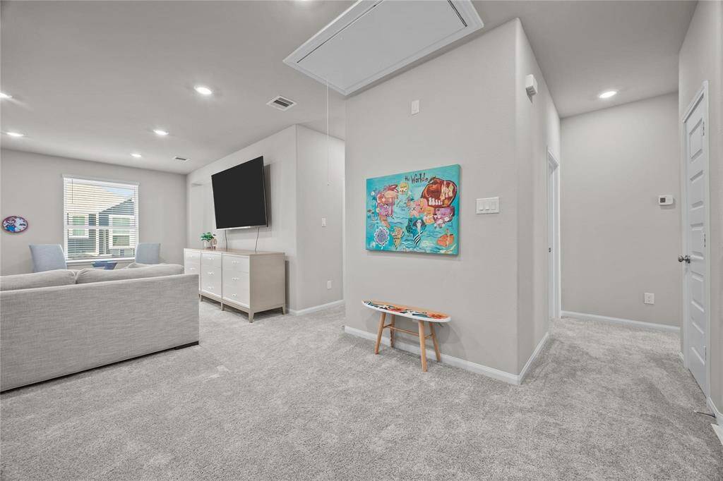 This photo showcases a modern, carpeted living area with neutral tones. It features a cozy seating arrangement, a large wall-mounted TV, and a console. The space is well-lit with recessed lighting and natural light from a window. The hallway leads to the primary bedroom, 3rd bedroom, and the laundry room.