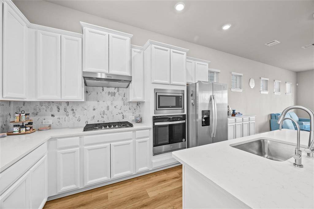 The gorgeous kitchen features sleek white cabinetry, a stainless steel refrigerator, and a built-in oven and microwave. It boasts a stylish backsplash, a gas cooktop, and a spacious island with a sink, perfect for meal prep and entertaining. Natural light streams through multiple windows, enhancing the bright, airy, and coastal feel.