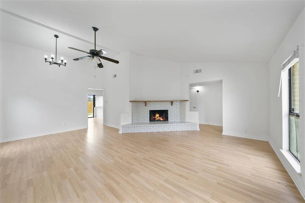 This spacious living room features light wood flooring, a cozy white brick fireplace, and high ceilings with a ceiling fan and chandelier. Large windows allow for ample natural light, creating an inviting atmosphere.
