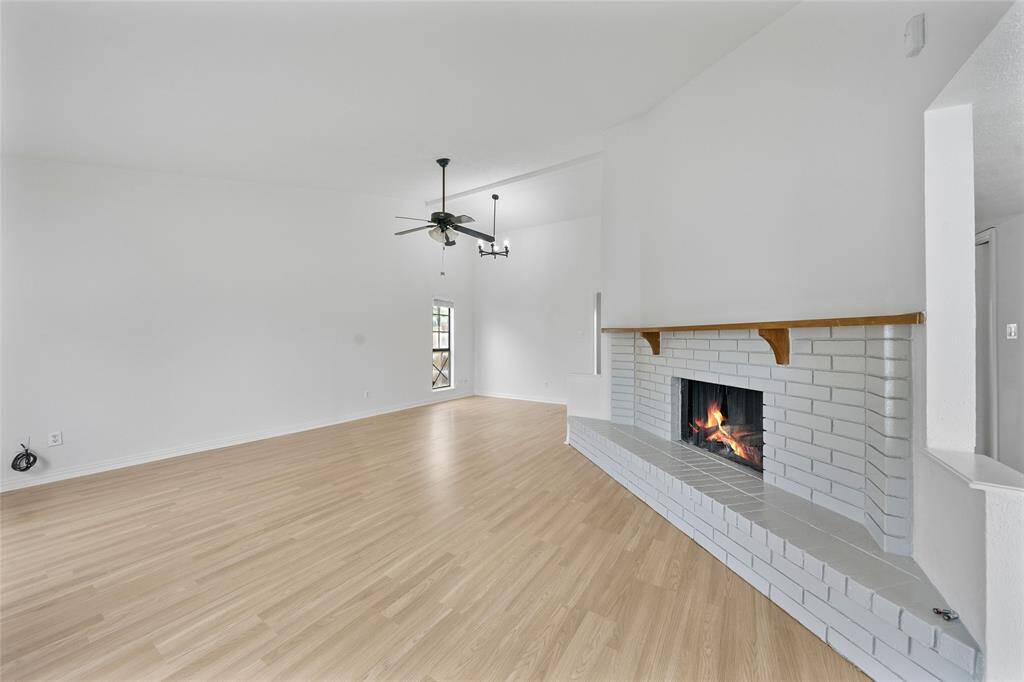 This spacious living room features a cozy white brick fireplace, light wood flooring, and a modern ceiling fan. The open design and neutral colors create a bright and inviting atmosphere.