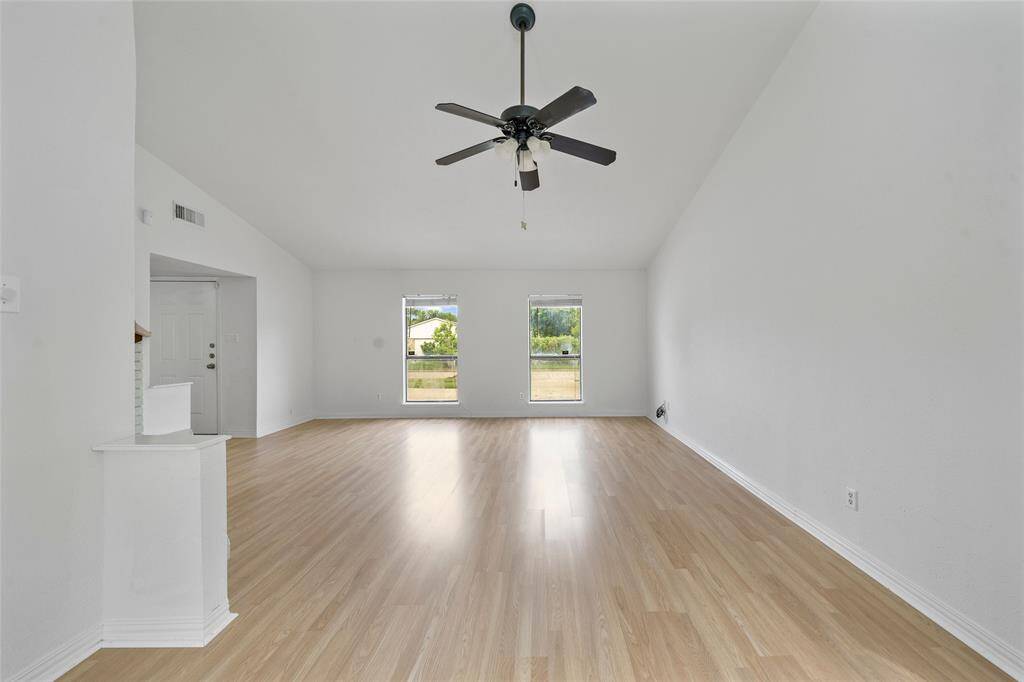 Bright, open living room with high ceilings, light wood flooring, and a ceiling fan. Two large windows provide natural light, enhancing the spacious feel.