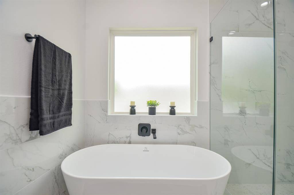 This bathroom features a modern freestanding bathtub with marble-style tiles, a frosted window for privacy, and sleek black fixtures.