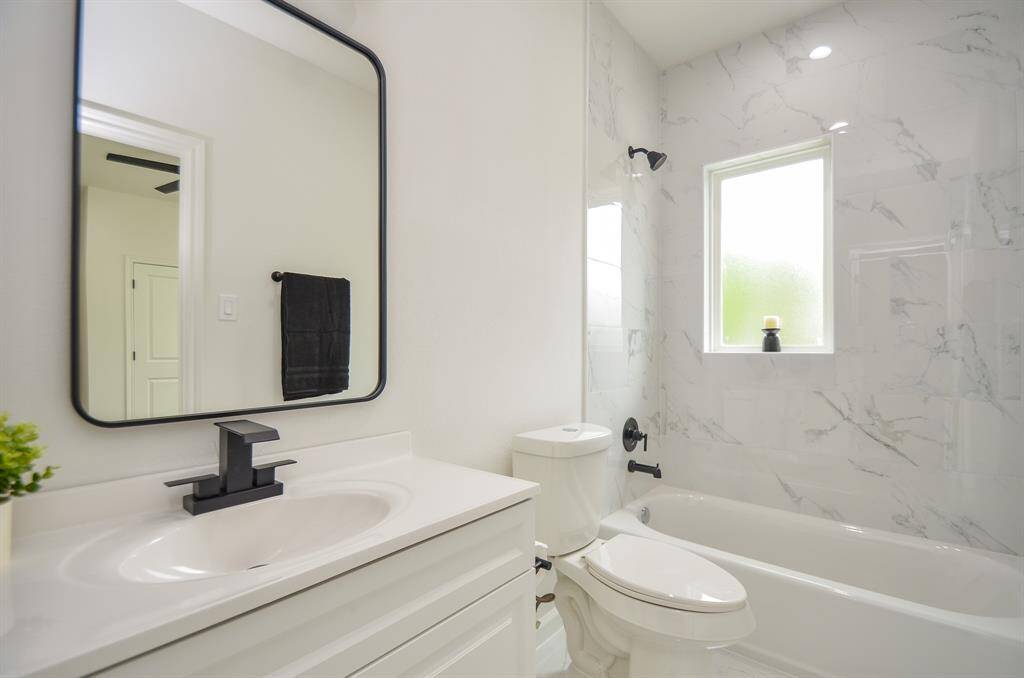 This bathroom features a sleek, modern design with a white vanity and black fixtures. It includes a bathtub with a marbled tile surround, a large mirror, and a frosted window for natural light.