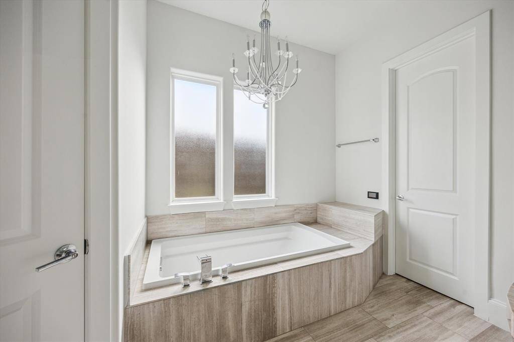 This primary bathroom features a large built-in soaking tub with tile surround, frosted windows for natural light, and a chandelier for a touch of elegance.