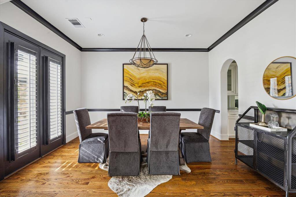 This formal dining room features rich hardwood flooring, crisp white walls, and bold black crown molding and trim. Double doors with shutters bring in natural light, and an arched doorway connects to the adjacent space.