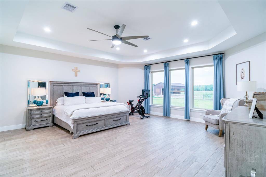 Bright and spacious Primary Bedroom with tile wood flooring. The room has ample natural light from three large windows with blue curtains, offering scenic views. Elegant furniture enhances the cozy, modern aesthetic.