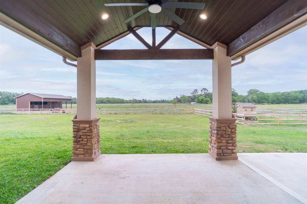 This photo showcases a spacious covered patio with stone pillars and a ceiling fan, overlooking a large grassy yard with a fenced area and a barn, ideal for country living.