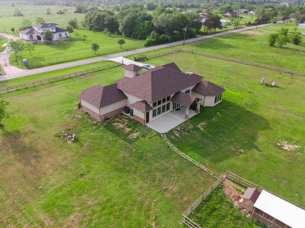 This aerial view showcases a spacious, single-story home with a modern design, surrounded by expansive green lawns. The property is fenced and located in a peaceful, rural setting with neighboring homes visible in the distance. Ideal for those seeking privacy and open space.