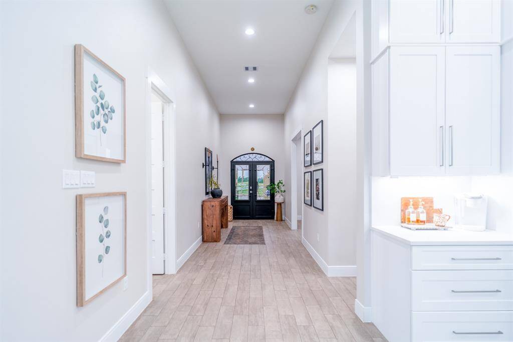 This photo showcases a bright, welcoming hallway with tile wood flooring and recessed lighting. It features modern decor with framed art, a stylish console table, and built-in cabinetry. The space leads to a decorative glass door, adding elegance and natural light.
